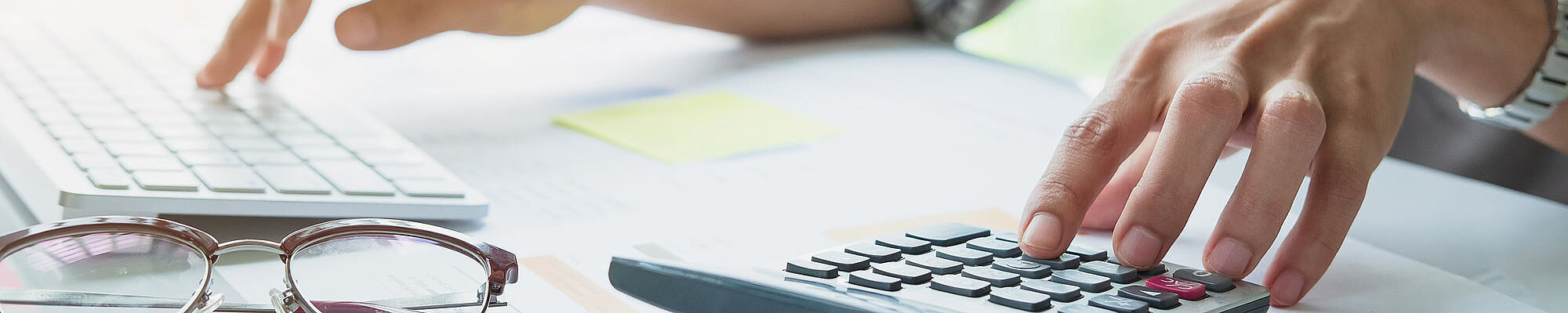 rechte Hand liegt auf einer Tastatur, linke Hand auf einem Taschenrechner