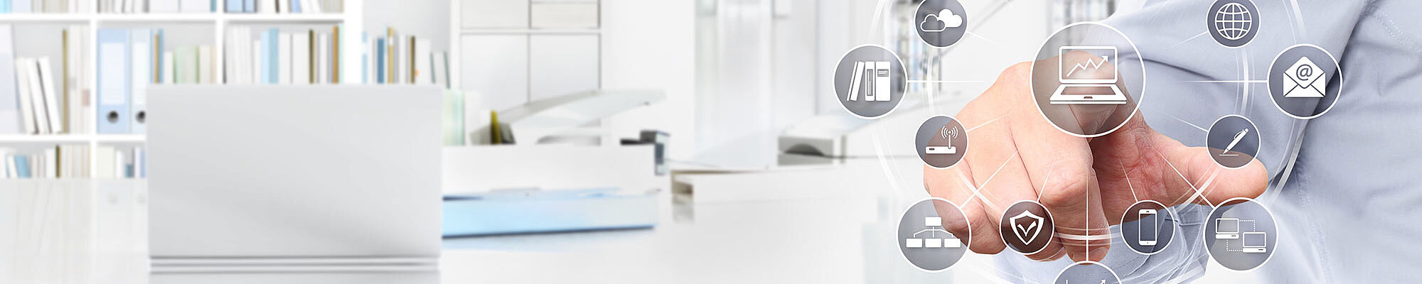 Büro mit Schreibtisch und Laptop. Im Vordergrund greift eine Hand zu einer grafischen Darstellung verschieder Icons aus dem IT-Bereich