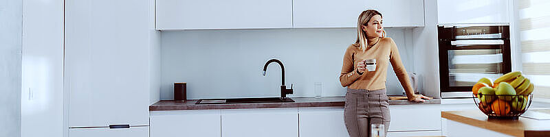 Teeküche Symbolbild Blonde Frau lehnt an heller Küchenzeile und hält eine Tasse in der Hand