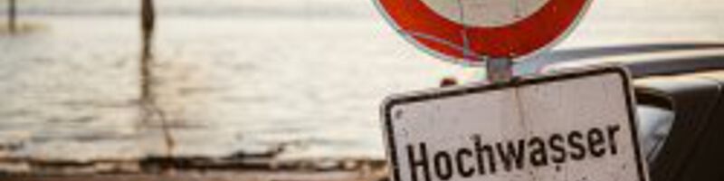 Hochwasser_Startseite_AdobeStock_321528897 Ein Schild wo Hochwasser darauf steht, steht vor einem See